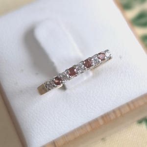May include: A delicate ring featuring alternating red and clear gemstones set in a gold band. The gemstones are small and round, creating a sparkling, elegant design. The ring is presented on a white surface.