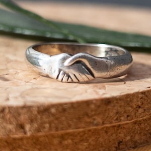May include: Silver ring featuring a handshake design. The ring is a band style with the clasped hands as the central focus. The metal has a slightly aged appearance. The ring is set against a natural wood surface.