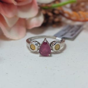 May include: A silver ring with a pear-shaped, deep red gemstone as the centerpiece. The ring features two smaller, round, yellow-toned stones on either side of the main stone. The ring is set against a soft, blurred background.