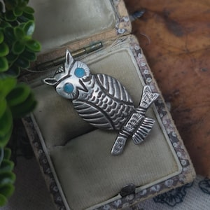 Puede incluir: Broche de búho plateado con ojos azul turquesa, posado en una rama. El búho está detallado con plumas grabadas y se exhibe dentro de un joyero vintage con un exterior estampado.