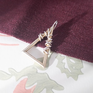 Può includere: Un ciondolo argentato a forma triangolare, con un design avvolto in filo metallico su un lato. Il ciondolo è posto su uno sfondo di tessuto bordeaux e una superficie a motivi.