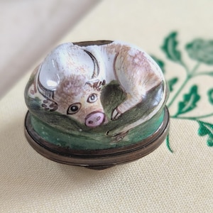Può includere: Piccola scatola rotonda per bigiotteria con un design bovino dipinto a mano. Il coperchio presenta una mucca a riposo nelle tonalità del bianco, beige e rosa, su una base verde. La scatola ha un bordo e una base in metallo color bronzo.