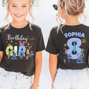 K&ouml;nnte beinhalten: Ein schwarzes T-Shirt mit den Worten "Birthday GIRL" in bunten Buchstaben und Zeichentrickfiguren. Auf der R&uuml;ckseite des Shirts steht der Name "SOPHIA" &uuml;ber einer gro&szlig;en "8" mit Zeichentrickfiguren. Das M&auml;dchen tr&auml;gt wei&szlig;e Shorts.