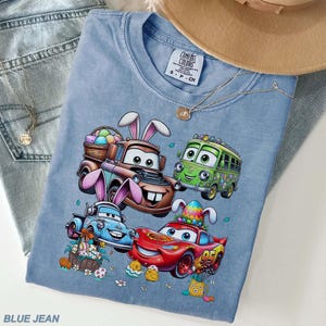 Pode incluir: Uma camiseta azul clara Comfort Colors com um gr&aacute;fico de personagens de Carros com orelhas de coelho e ovos de P&aacute;scoa. A camisa &eacute; combinada com jeans azuis, um colar e um chap&eacute;u bege. O texto "BLUE JEAN" est&aacute; na parte inferior.