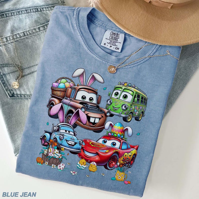 Pode incluir: Uma camiseta azul clara Comfort Colors com um gr&aacute;fico de personagens de Carros com orelhas de coelho e ovos de P&aacute;scoa. A camisa &eacute; combinada com jeans azuis, um colar e um chap&eacute;u bege. O texto "BLUE JEAN" est&aacute; na parte inferior.