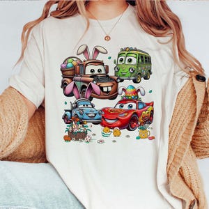 Pode incluir: Uma camiseta creme com um gr&aacute;fico colorido de carros animados decorados para a P&aacute;scoa. Os carros incluem um caminh&atilde;o marrom com orelhas de coelho, um &ocirc;nibus verde, um caminh&atilde;o azul e um carro de corrida vermelho. Ovos e pintinhos de P&aacute;scoa tamb&eacute;m s&atilde;o retratados.