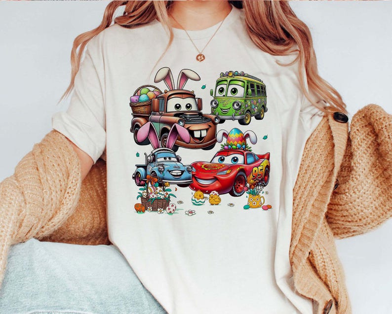 Pode incluir: Uma camiseta creme com um gr&aacute;fico colorido de carros animados decorados para a P&aacute;scoa. Os carros incluem um caminh&atilde;o marrom com orelhas de coelho, um &ocirc;nibus verde, um caminh&atilde;o azul e um carro de corrida vermelho. Ovos e pintinhos de P&aacute;scoa tamb&eacute;m s&atilde;o retratados.