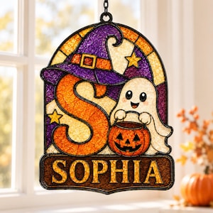 May include: A stained glass-style hanging decoration featuring a large orange letter S, a purple witch hat, and a white ghost holding an orange jack-o'-lantern bucket. The base displays the name SOPHIA in yellow letters on a dark brown background.