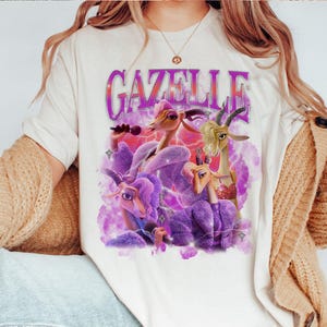Puede incluir: Camiseta color crema con la palabra "GAZELLE" en fuente morada estilizada. El gr&aacute;fico muestra cabras de dibujos animados en morado y beige, con un fondo de rayos rosas y morados.