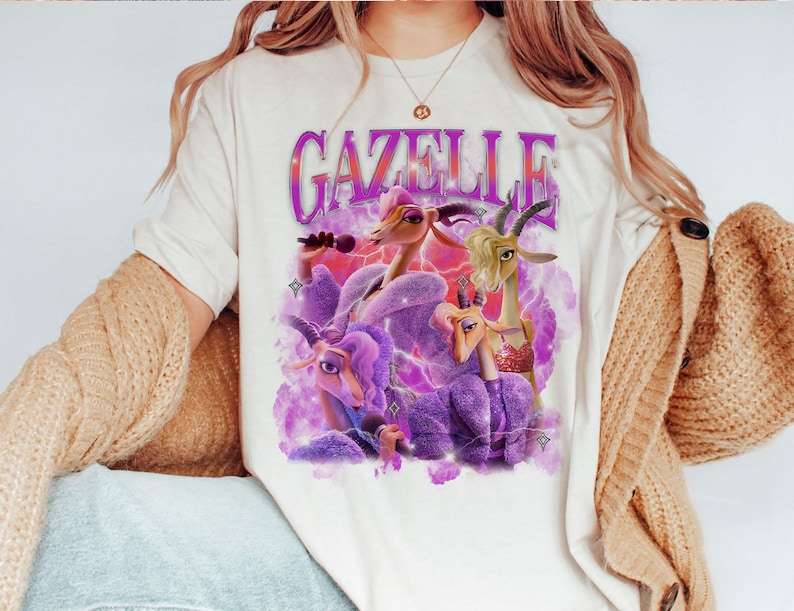 Puede incluir: Camiseta color crema con la palabra "GAZELLE" en fuente morada estilizada. El gr&aacute;fico muestra cabras de dibujos animados en morado y beige, con un fondo de rayos rosas y morados.