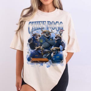 Puede incluir: Camiseta beige con un gr&aacute;fico del Jefe Bogo y otros personajes de Zootopia. El dise&ntilde;o incluye el texto "CHIEF BOGO" en una fuente azul estilizada. La imagen representa a los personajes con uniformes de polic&iacute;a.