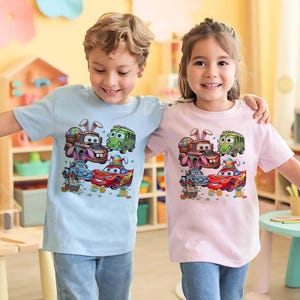 Pode incluir: Duas crian&ccedil;as vestindo camisetas azul claro e rosa. Cada camisa apresenta um gr&aacute;fico colorido de carros de desenho animado decorados com ovos de P&aacute;scoa e orelhas de coelho. As crian&ccedil;as est&atilde;o sorrindo e se abra&ccedil;ando.