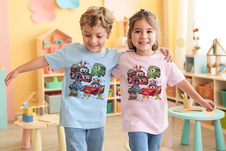 Pode incluir: Duas crian&ccedil;as vestindo camisetas azul claro e rosa. Cada camisa apresenta um gr&aacute;fico colorido de carros de desenho animado decorados com ovos de P&aacute;scoa e orelhas de coelho. As crian&ccedil;as est&atilde;o sorrindo e se abra&ccedil;ando.