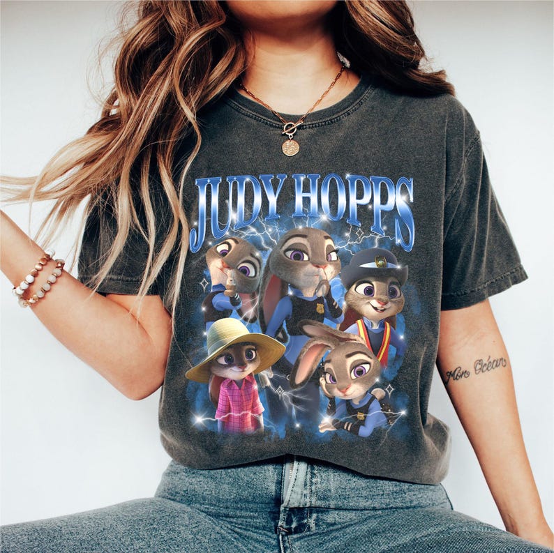 Puede incluir: Camiseta gris oscuro con el texto azul "JUDY HOPPS" y un gr&aacute;fico del personaje Judy Hopps de la pel&iacute;cula Zootr&oacute;polis. El gr&aacute;fico muestra a Judy en varias poses y atuendos, con un fondo de rayos azules.