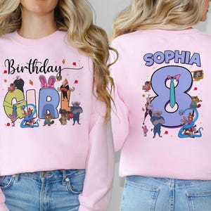 K&ouml;nnte beinhalten: Hellrosa Sweatshirt mit Aufdruck "Birthday GIRL" auf der Vorderseite und "SOPHIA 8" auf der R&uuml;ckseite. Das Design zeigt Cartoon-Figuren und den Schriftzug "Birthday" in Schreibschrift. Auf dem R&uuml;cken befindet sich eine gro&szlig;e 8.