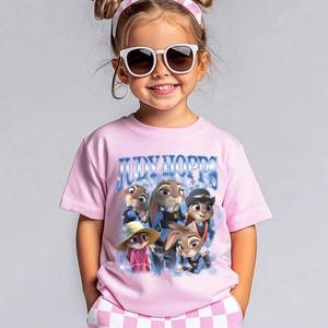 Puede incluir: Una ni&ntilde;a peque&ntilde;a con una camiseta rosa con la frase "JUDY HOPPS" y un gr&aacute;fico de personajes de la pel&iacute;cula Zootopia. Tambi&eacute;n lleva gafas de sol y una diadema rosa y blanca.