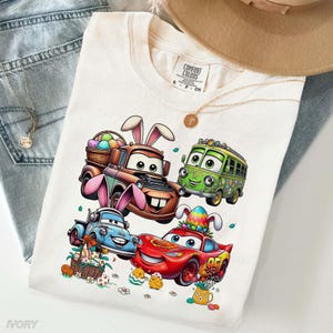 Pode incluir: Camiseta Comfort Colors marfim com um gr&aacute;fico colorido de carros animados com orelhas de coelhinho da P&aacute;scoa e ovos. Os carros s&atilde;o vermelhos, azuis, verdes e castanhos. A camiseta &eacute; combinada com jeans azuis e um colar dourado.