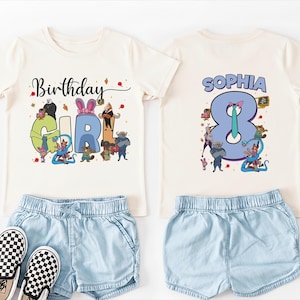 K&ouml;nnte beinhalten: Ein cremefarbenes Geburtstags-T-Shirt mit den Worten "Birthday GIRL" in bunten Buchstaben und Zeichentrickfiguren. Auf der R&uuml;ckseite des Shirts steht der Name "SOPHIA" &uuml;ber der Zahl "8". Kombiniert mit hellblauen Jeansshorts und karierten Slip-On-Schuhen.