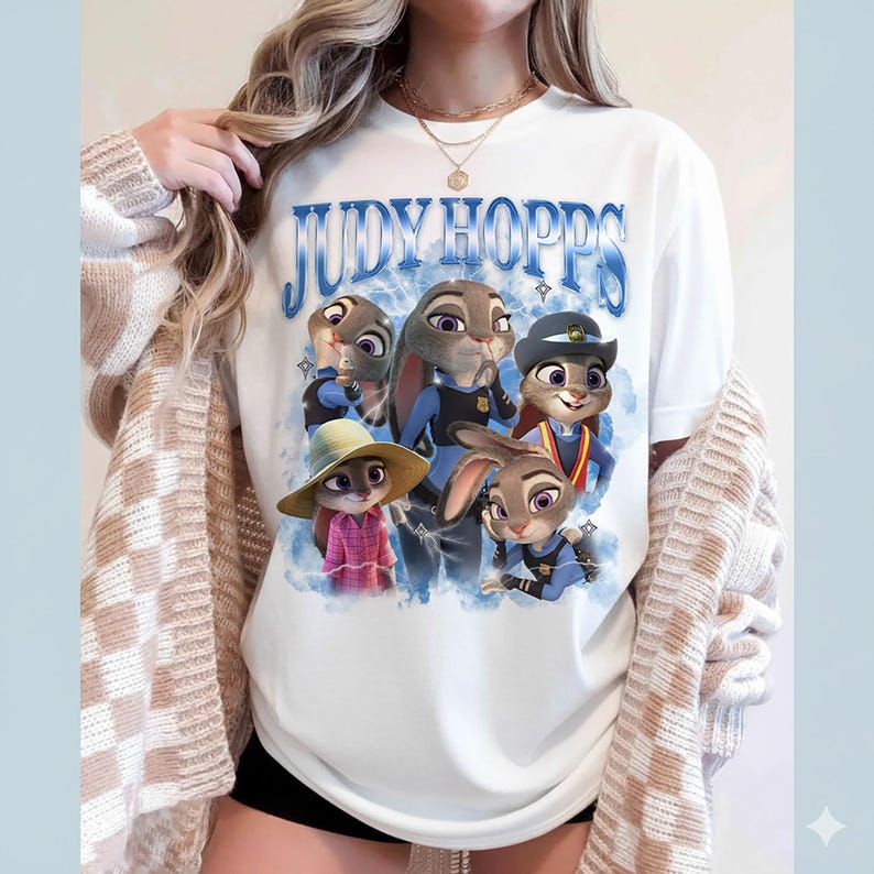 Puede incluir: Camiseta blanca con el texto azul "JUDY HOPPS" y un gr&aacute;fico de conejos de dibujos animados con uniformes de polic&iacute;a y ropa de civil. Los conejos est&aacute;n en varias poses sobre un fondo azul nublado.