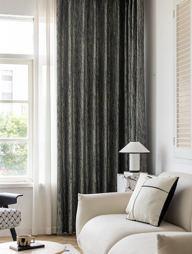 May include: Dark gray and beige patterned curtain hanging in front of a window. The curtain has vertical lines and is next to a sheer white curtain. A white lamp sits on a marble table next to a white sofa with a black and white pillow.