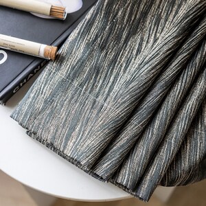 May include: Close-up of folded fabric with a textured pattern in shades of gray and beige. The fabric is draped over a white surface, with a book and incense sticks visible in the background. The fabric's design resembles brushstrokes.