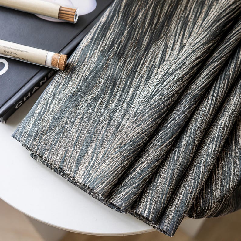 May include: Close-up of folded fabric with a textured pattern in shades of gray and beige. The fabric is draped over a white surface, with a book and incense sticks visible in the background. The fabric's design resembles brushstrokes.