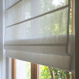 May include: A light-colored Roman shade made of a textured fabric, partially covering a window. The shade is in a neutral tone, with horizontal folds. The window frame is visible, and the background shows a view of green foliage.