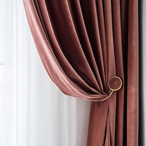 May include: Close-up of a luxurious, dusty rose velvet curtain. The curtain is held back with a gold-colored circular tieback. Sheer white curtains are visible behind the velvet curtain.