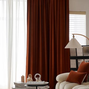 May include: Interior shot with rust-coloured velvet curtains and white sheer curtains. A white sofa with rust-coloured cushions is in the foreground. A side table displays ornaments. A lamp and a bookshelf are also visible.