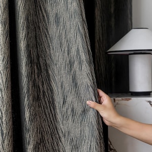 May include: Dark gray and gold patterned curtain fabric. The fabric has a textured, woven appearance with vertical lines of gold against a dark gray background. The curtain is held open, revealing the pattern and texture.