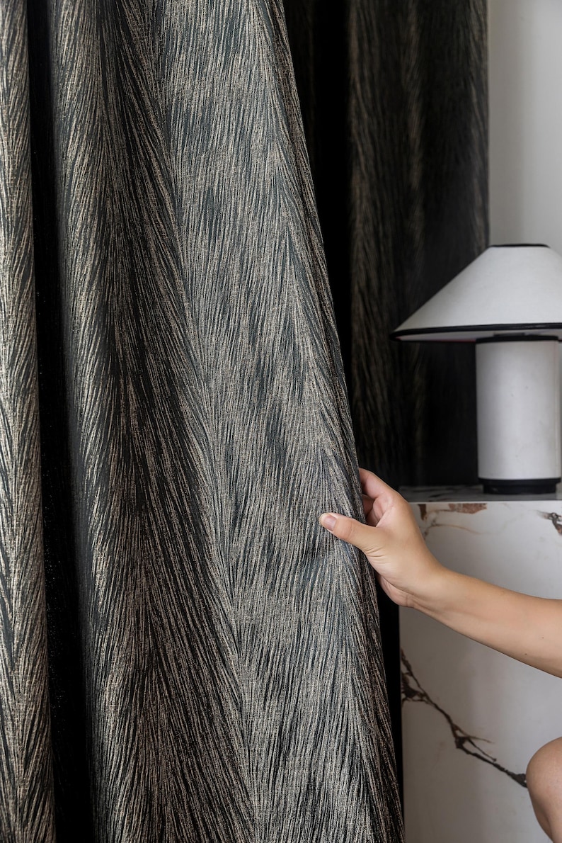 May include: Dark gray and gold patterned curtain fabric. The fabric has a textured, woven appearance with vertical lines of gold against a dark gray background. The curtain is held open, revealing the pattern and texture.