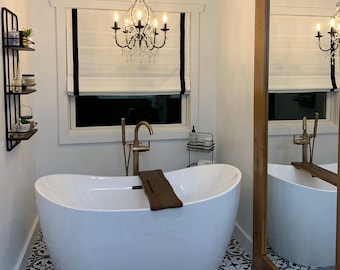 Puede incluir: Un baño moderno con una bañera blanca de forma ovalada. Una bandeja de madera descansa sobre la bañera. La habitación cuenta con una lámpara de araña negra, un espejo grande y un suelo de baldosas estampadas. Una estantería de metal negro contiene artículos de aseo. La ventana tiene persianas blancas con ribete negro.