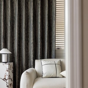 May include: A modern living room scene featuring a textured, dark gray curtain. A white sofa with a decorative pillow and a marble side table with a lamp are in the foreground. The room has a minimalist aesthetic.