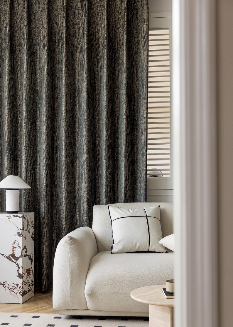 May include: A modern living room scene featuring a textured, dark gray curtain. A white sofa with a decorative pillow and a marble side table with a lamp are in the foreground. The room has a minimalist aesthetic.