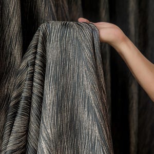 May include: Dark gray fabric with vertical, textured lines in a lighter, metallic gold color. The fabric is held up, showing the drape and texture of the material. The fabric appears to be a curtain or drapery material.