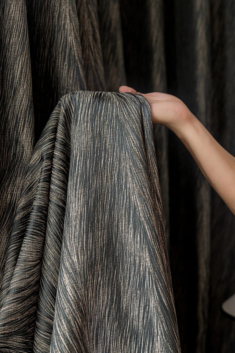 May include: Dark gray fabric with vertical, textured lines in a lighter, metallic gold color. The fabric is held up, showing the drape and texture of the material. The fabric appears to be a curtain or drapery material.