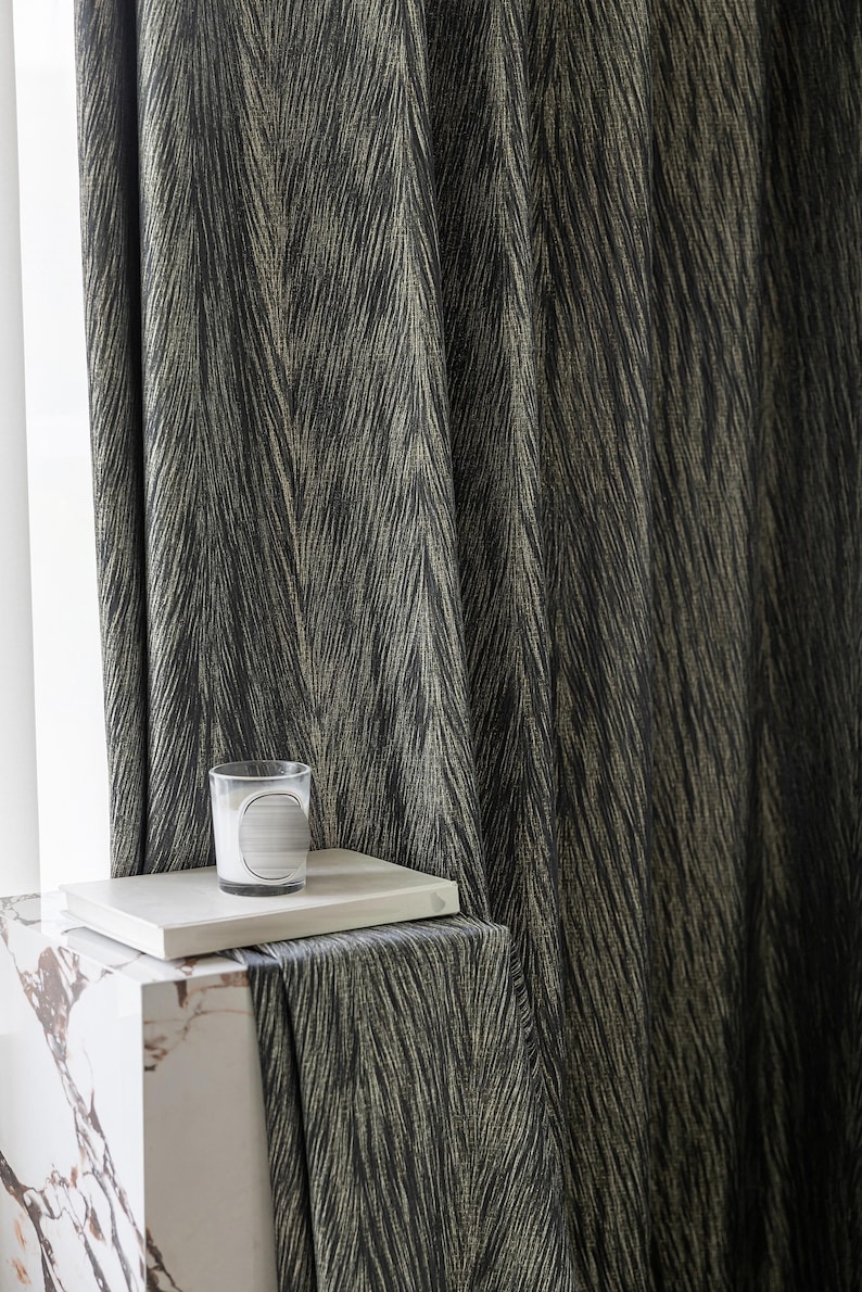May include: Dark gray and gold patterned curtain fabric. A small glass candle holder with a white label sits on a white book. The book rests on a white marble block. The curtain fabric has a textured, diagonal stripe design.