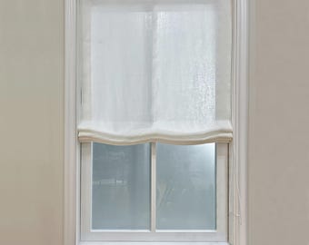 Puede incluir: Una cortina romana blanca está parcialmente bajada en una ventana con marco blanco. La cortina está hecha de una tela transparente y de color claro. El marco de la ventana y la pared circundante están pintados de un color neutro.