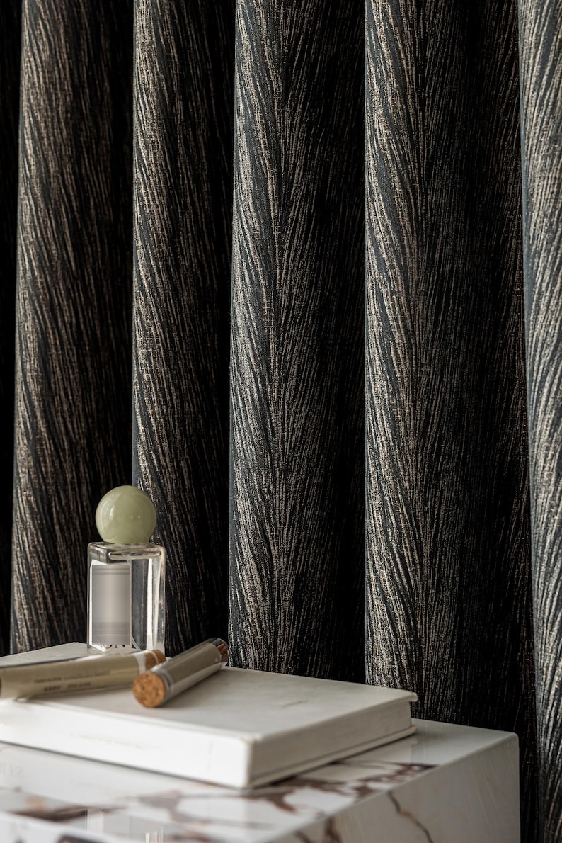 May include: Dark, textured curtains with a feather-like pattern in black and gold tones. A clear perfume bottle with a green orb-shaped stopper sits on a white book, with two small glass vials with cork stoppers.