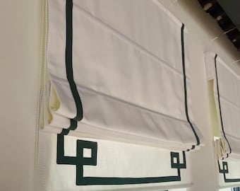 May include: White Roman shades with dark green trim and a geometric design at the bottom. The shades are hanging in front of a window. The shades are made of a light-colored fabric and have a clean, modern look.