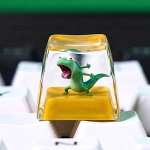 May include: A novelty keycap with a green lizard figurine inside a clear, rectangular casing, set on a yellow base. The keycap sits on a white keyboard, with a green backdrop.