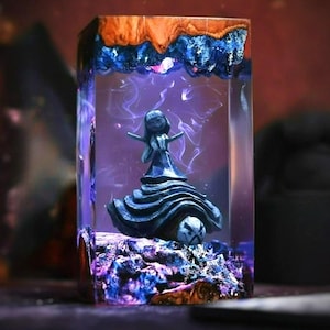 May include: A rectangular resin sculpture featuring a dark blue figurine with outstretched arms, encased in clear resin. The top has a wood grain appearance with blue accents. The base has a rocky texture with purple and blue hues.