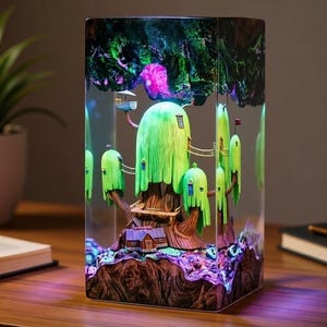 May include: A decorative lamp featuring a whimsical treehouse design encased in a clear rectangular block. The treehouse has green, yellow, and brown elements, with small houses and bridges. The lamp emits a colorful glow, creating a fantasy scene.