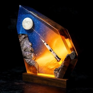 May include: A unique resin sculpture featuring a rocket launching into space, with a full moon and starry sky. The sculpture is encased in a clear, geometric form, set on a wooden base, and illuminated from within.