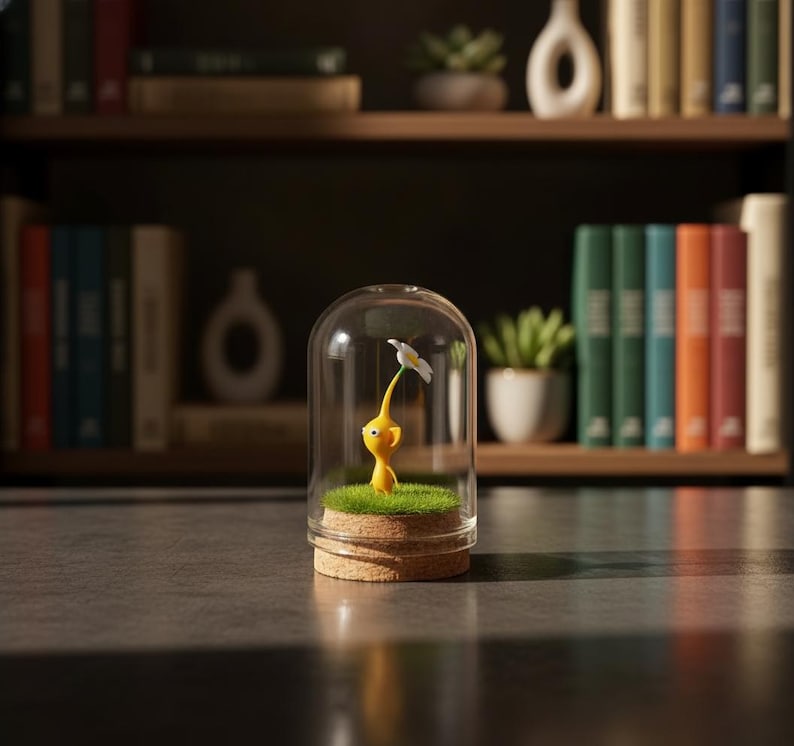 May include: A yellow figurine of a character holding a white flower, encased in a glass dome. The figurine stands on a bed of green grass, atop a cork base. The background features a bookshelf with books and plants.