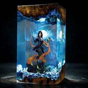 Könnte beinhalten: Rechteckige Kunstharzskulptur mit einer weiblichen Figur in dunkler Kleidung, die auf einem detaillierten Sockel mit orangefarbenen und schwarzen Akzenten steht. Die Figur ist in einem transparenten Kunstharzblock mit ätherischen blauen und weißen Lichteffekten und einem Holzrahmen eingeschlossen.