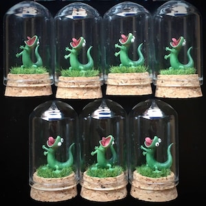 May include: A collection of miniature terrariums, each featuring a green lizard figurine with an open mouth, set on a bed of green faux grass. The lizards are enclosed in clear glass domes atop cork bases. The background is black.