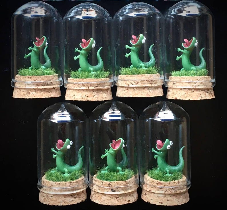 May include: A collection of miniature terrariums, each featuring a green lizard figurine with an open mouth, set on a bed of green faux grass. The lizards are enclosed in clear glass domes atop cork bases. The background is black.