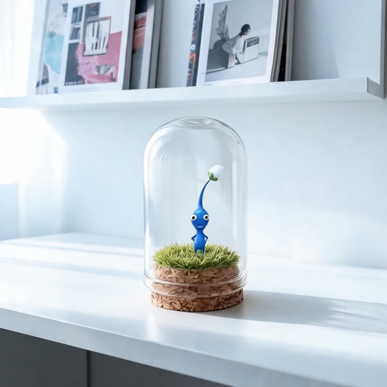 May include: A blue Pikmin figure with a white flower, enclosed in a glass cloche. The figure stands on a bed of green faux grass, set on a cork base. The cloche is displayed on a white shelf.
