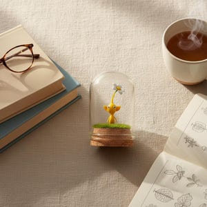May include: A yellow figurine with a white flower under a glass dome. The figurine sits on a patch of green grass atop a cork base. A cup of tea, books, and eyeglasses are also in the image.